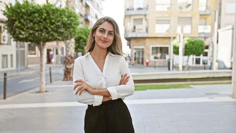 Cheerful, confident young blonde woman enjoying the sunny city atmosphere, standing on a street with a joyful smile and relaxed arms crossed gesture