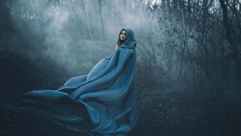 A big, beautiful woman in a blue raincoat, walks in a fog. Background dark forest, bare trees. Creative colors