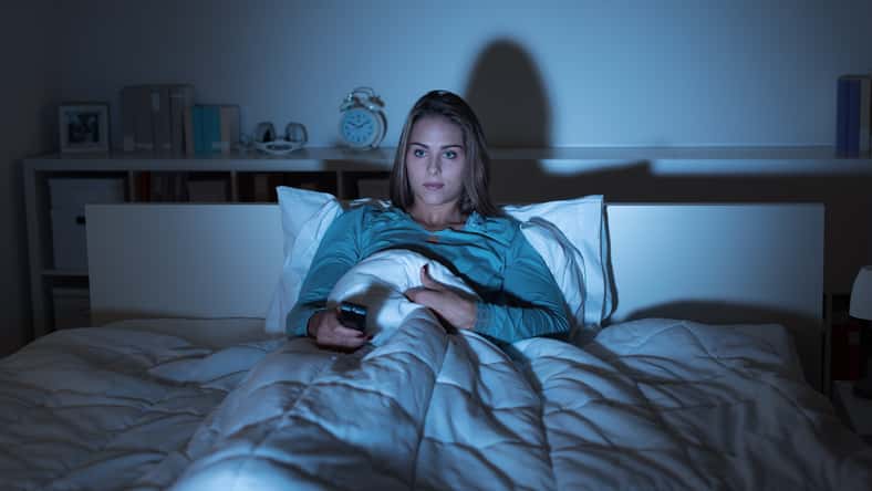 Young woman relaxing in bed late at night and watching tv, she is holding a remote control