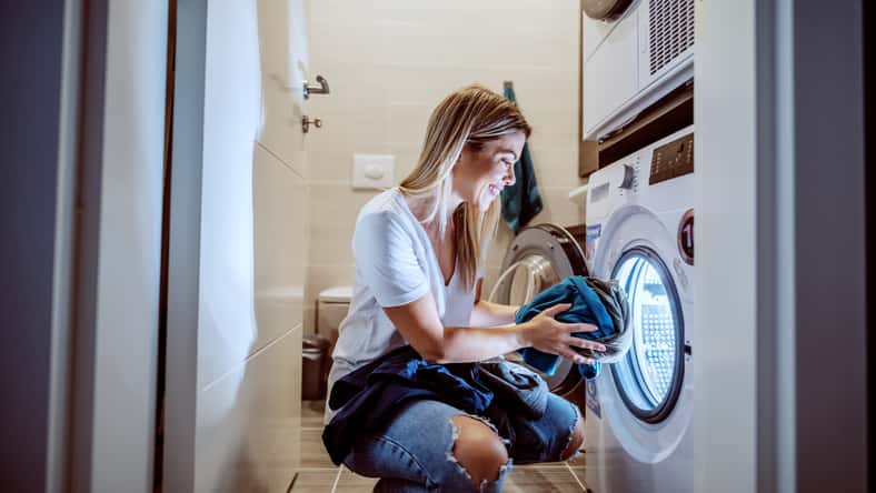 Smiling caucasian blond worthy housewife dressed casual crouching in bathroom and loading washing machine with dirty clothes in late hours.
