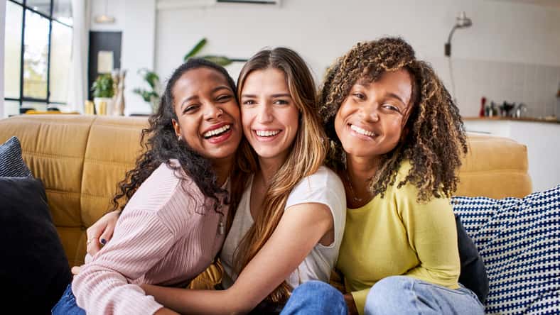 Portrait of three attractive female room mates looking smiling at camera. Group of cute cheerful girls hugging sitting on couch at home. Happy friends posing for photo indoors. Generation z and rent.