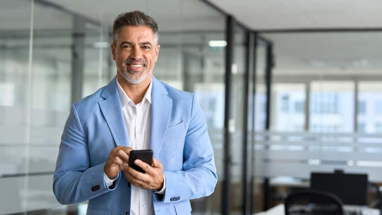 Happy mature business man investor wearing suit standing in office using mobile phone. 50 year old smiling businessman professional holding smartphone looking at camera at work. Portrait, copy space.