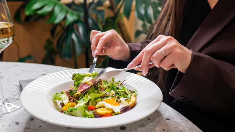 A person elegantly dining, using a fork and knife to enjoy a fresh salad. A glass of white wine is nearby, set in a stylish, modern indoor environment with greenery in the background.