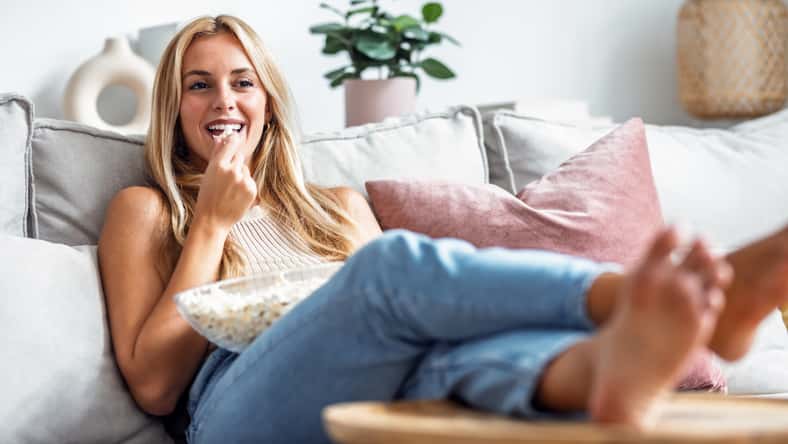 Shot of laughing young woman watching a movie on tv while eating popcorn lying on the couch at home