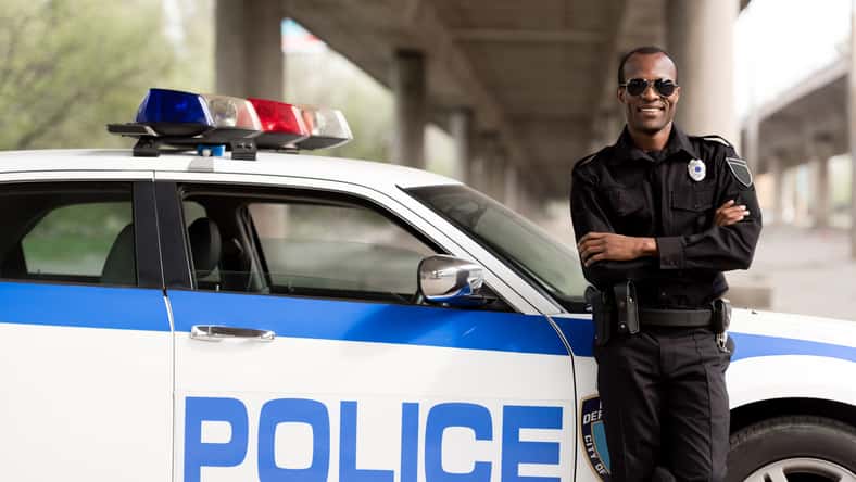 handsome african american police officer with crossed arms leaning back on car and looking at camera