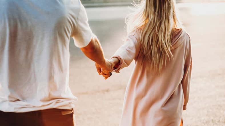 Crop back view of casual couple holding hands and walking on street in sunshine
