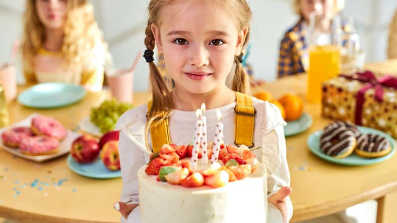 girl with birthday cake in hands, she looks at camera, celebrating with friends, kids behind table in the background