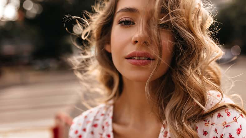 Close-up of mysterious caucasian young lady with imposing facial expression looking at camera. Gorgeous blonde looks at camera wearing white blouse. Happiness summer lifestyle concept.