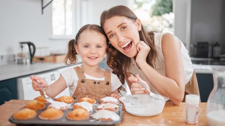 Baking, portrait and decorating with mother, daughter and learning cooking in home kitchen. Food, kids and help with girl and mom happy with muffins, cupcakes and icing results for bonding together.