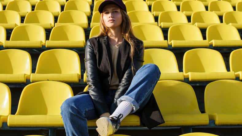 Imagen de estilo de vida. Hermosa chica en un cálido día soleado en la ciudad del estadio. Mujer joven de pelo largo sentado en las gradas con el pie en el asiento y nos mira. Moda de la calle de verano.