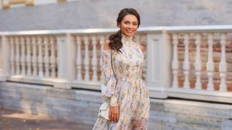 Pretty smiling young woman with long brunette hair wearing fashionable floral dress standing on street against balustrade on background. Happy female model in elegant stylish outfit posing outdoors.
