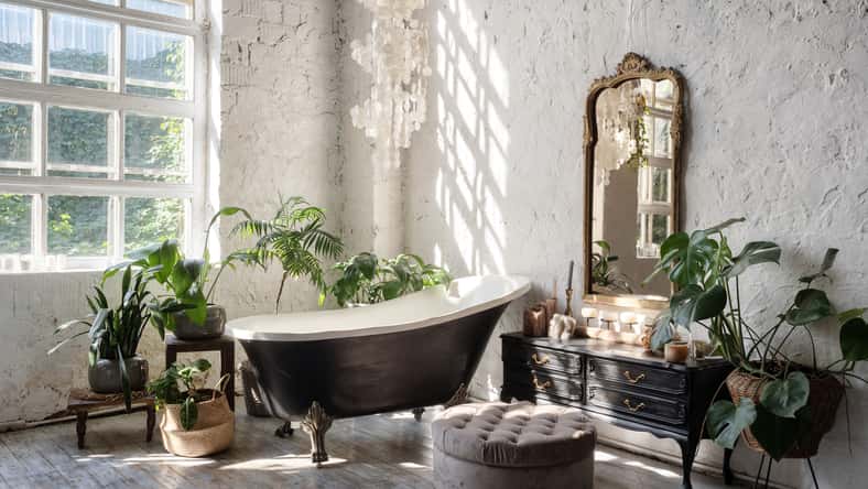 Side view of white cozy bathroom with black tub, wooden classic commode, mirror, decor, plants and interior design in boho chic style. Concept of comfortable and decorated room at home