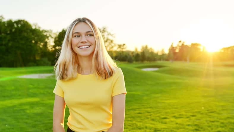 Outdoor portrait of smiling teenage girl 16, 17 years old in yellow T-shirt, on green sunny lawn. Sunset, grass background for copy space
