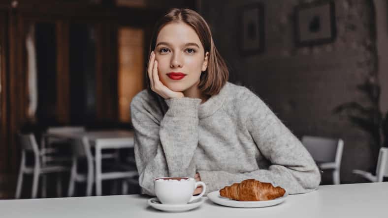Brown-eyed girl in woolen sweater leaned on white table in cafe and looking at camera. Photo of woman with red lips ordering coffee and croissant