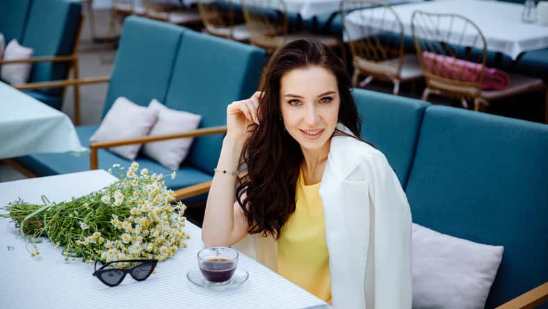 Portrait of a beautiful smiling girl sitting at table in summer cafe in the street of an old European city with bouquet of flowers. Young lady in restaurant. Europe vacation, romantic dinner.