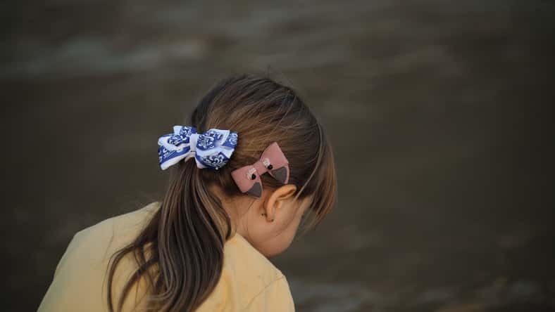 Hairstyle of Caucasian little girl close up - brown hair gathered in tail and two bows blue and pink. Beautiful hairstyle of child is little side view from behind.