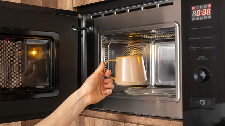 A person's hand reaches into a microwave to place inside a ceramic mug with cold beverage to heat coffee at home or work