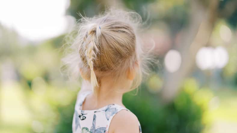 Little girl with flowing hair stands near a tree and points to it. Back view. High quality photo