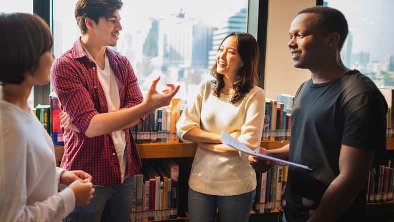 Group of diverse young adults in a library, discussing and smiling. Casual attire, diverse ethnicities, engaging in conversation, standing by bookshelves. Diverse high school students talking.