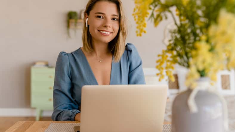 young pretty stylish woman working remote at home at table workplace, student education online, fashion blog trend, wearing blouse, smiling looking in camera, using earpods