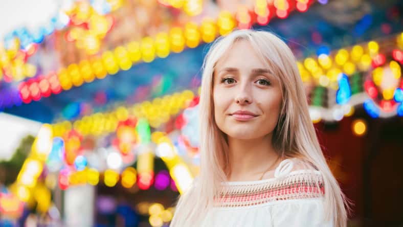 Young attractive blonde girl standing near carousel in amusement park. Beautiful portrait of woman with long hair.