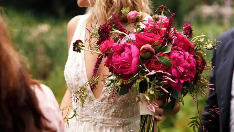 Pretty bride holds rich dark pink bouquet of peonies in her arm