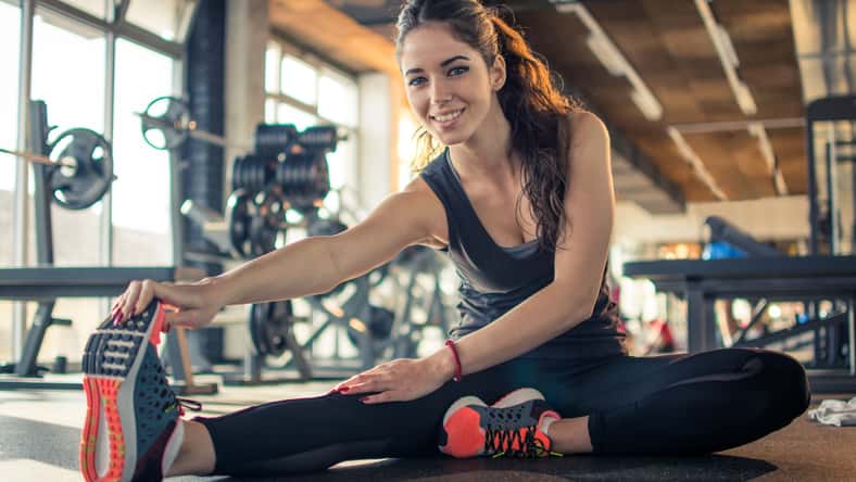 Smiling fit girl stretching legs in gym.