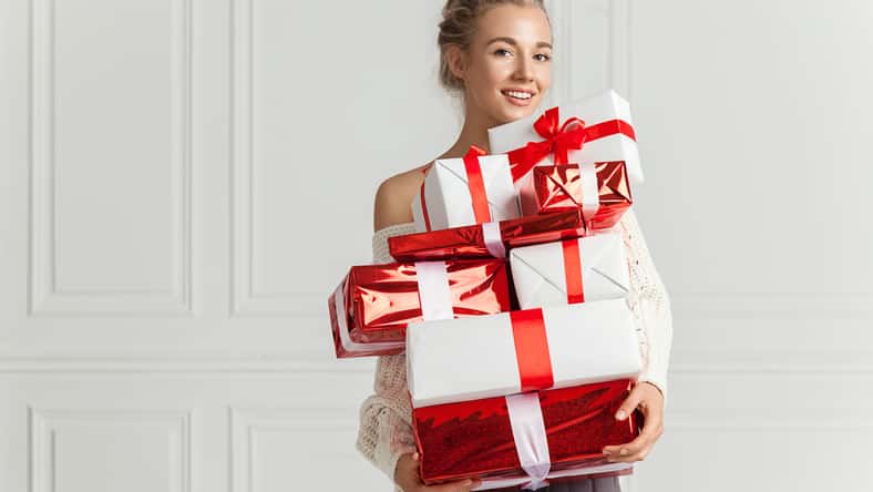Blond hair woman in white sweater holding a lot of presents over white wall background indoor. Smiling woman holding paper gift boxes red color and looks at camera. Toothy smiling woman's face.
