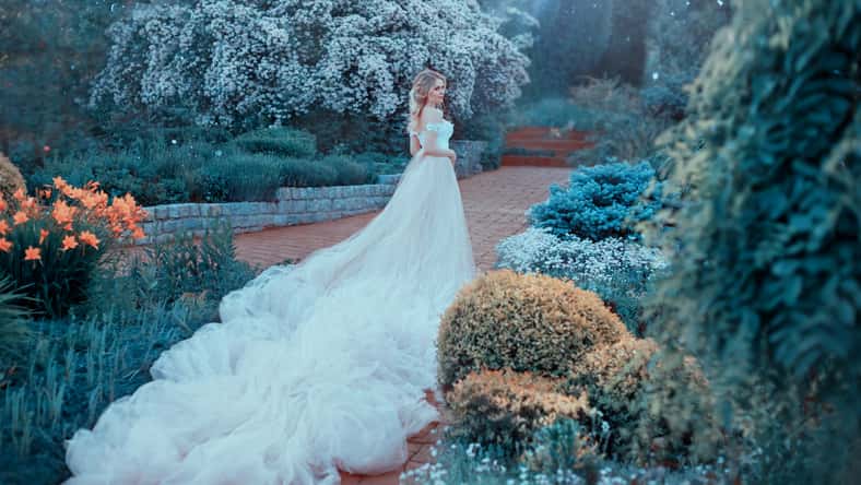 Blonde, with a beautiful elegant hairdo, walks in a fabulous blooming garden. Princess in a luxurious light pink dress with an incredibly long train goes to the next. Photo from the back. Art photo.
