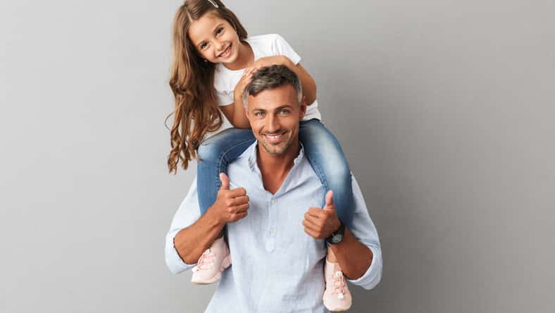 Portrait of adorable family, daughter smiling and sitting on the neck of her father 30s isolated over gray background