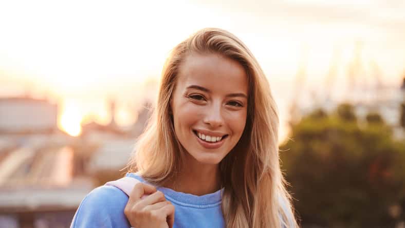 Portrait of a smiling young girl with backpack standing outdoors during sunset
