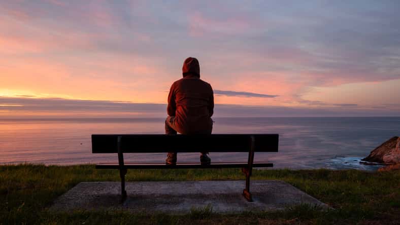 Lonely man sits alone on the rocky coast and enjoying sunset. View over rocky cliff to ocean horizon