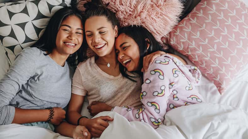 Three girls laughing and having fun while lying on bed together during a sleepover. Top view of young girls having a sleepover lying on bed and laughing.