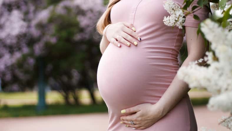 pregnant blonde girl in pink dress with bare shoulders holding hands belly with long hair on the background of lilacs and trees with a big belly on the eighth month of pregnancy close-up