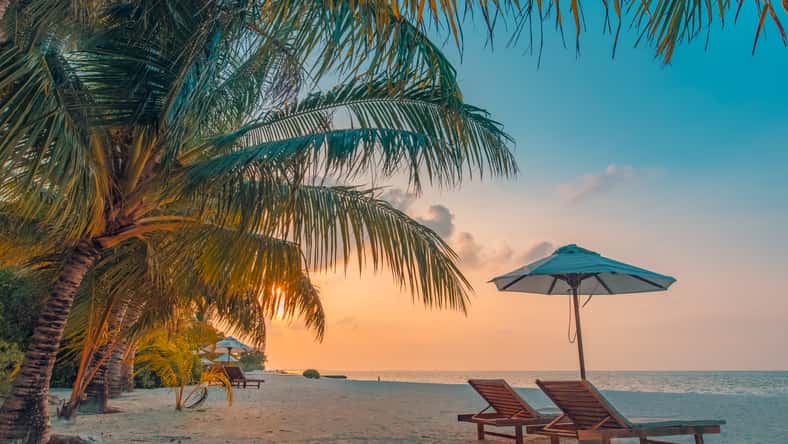 Beautiful tropical sunset scenery, two sun beds, loungers, umbrella under palm tree. White sand, sea view with horizon, colorful twilight sky, calmness and relaxation. Inspirational beach resort hotel