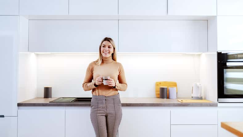 Attractive fashionable Caucasian smiling blonde woman leaning on kitchen counter and holding mug with coffee. Apartment interior.