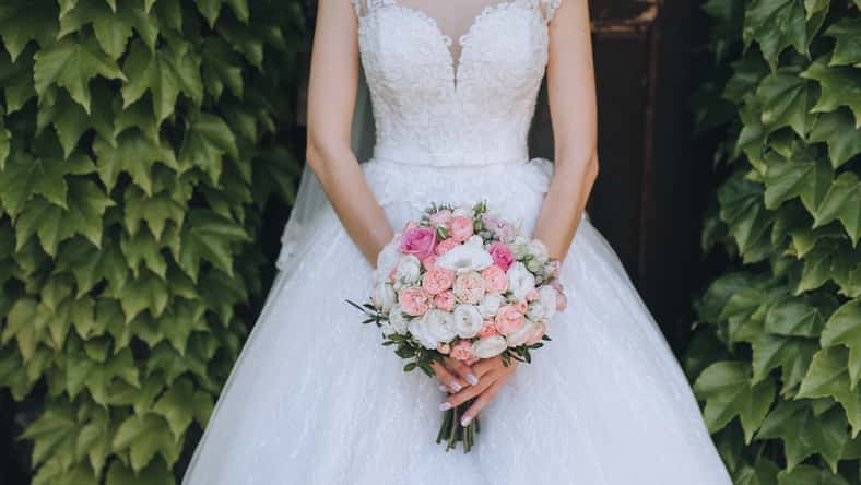The bride in a white dress holds a wedding bouquet of roses in her hands. Closeup portrait of fresh flowers on nature, against a background of green foliage. Photography, concept.