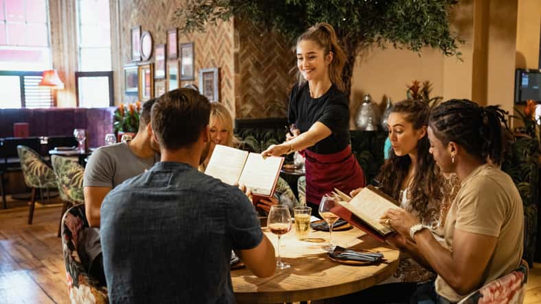 A multi-ethnic group of friends looking at menus together in a restaurant, which have been given to them by a waitress.