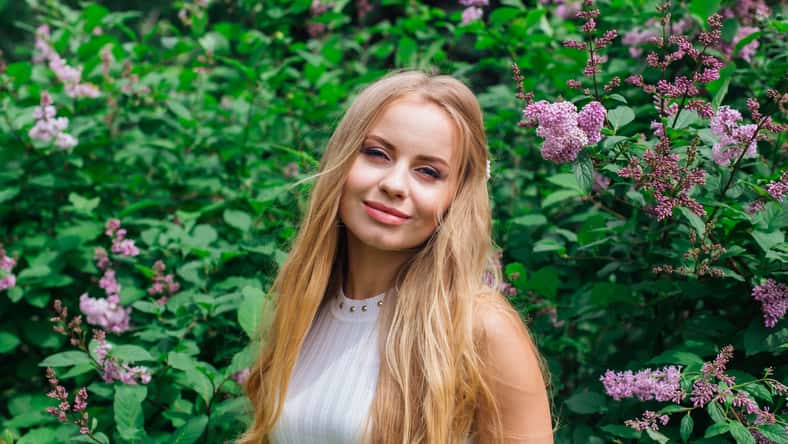 Spring portrait of a charming blond woman wearing beautiful white dress standing next to blooming purple lilac bush.