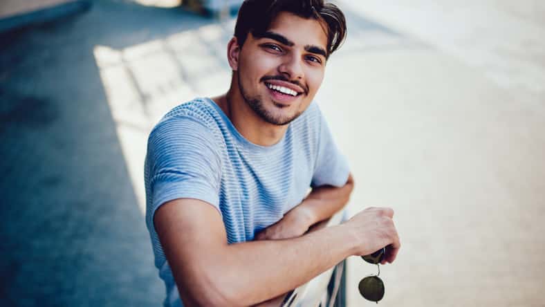 Portrait of cheerful bearded hipster guy waiting for meeting on stairs enjoying spring sunny day, happy young man with brown hair in casual outfit smiling at camera spending time outdoors