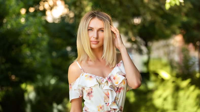 Attractive young blonde woman in a light summer dress with flowers, woman enjoying her time outdoors in a summer green park with the sunset in the background.