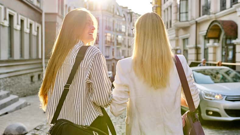 Two young women with laptop bags walking along street of sunset city, back view. Urban style background, female university students, colleagues office workers women