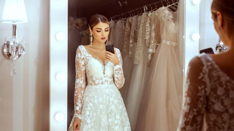 Portrait of a young bride in a wedding boutique. She takes pictures of herself in the mirror on her phone.