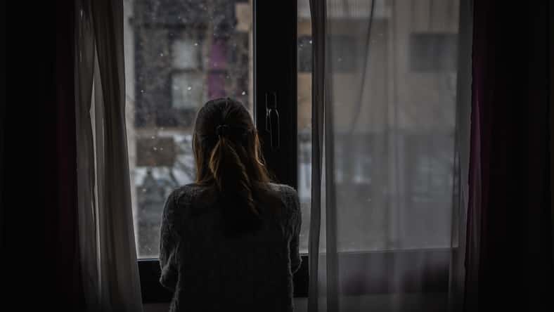 Young woman looks out a window between the curtains while it is snowing.