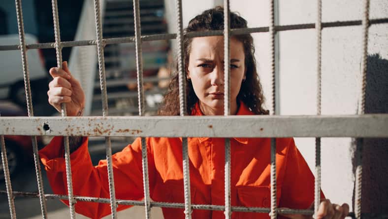 Young woman in orange suit behind jail bars. Female in colorful overalls portrait. Law and justice concept