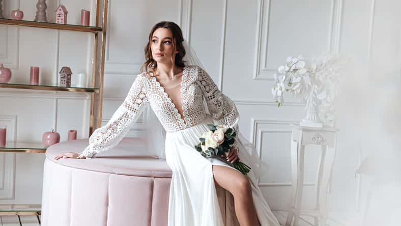An attractive young bride in a beautiful boho wedding dress posing with a bouquet in a bright room. Wedding photo session, bride's morning.