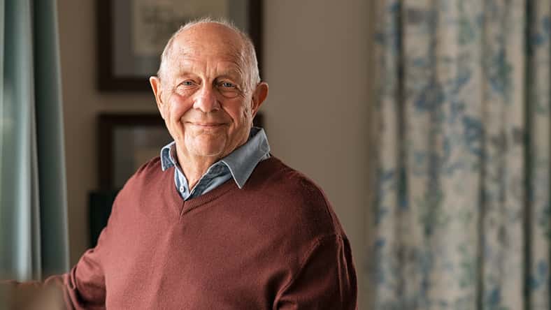 Portrait of happy retired senior man standing at home near window. Satisfied old man in casual clothes looking at camera and smiling while standing near the window. Positive and confident elderly enjoy his retirement at care facility.