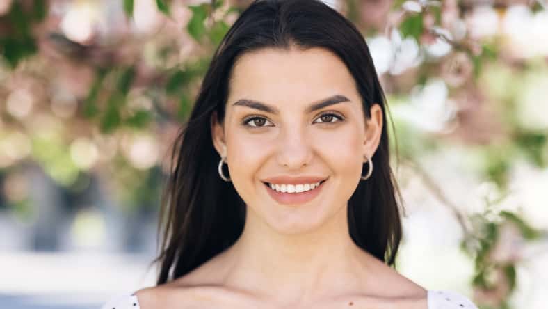 Portrait of a Gorgeous Dark Haired Woman Smiling Wearing Sammer Dress Charmingly while Standing at park on a background of sakura trees. Happy Young Woman Enjoys Life
