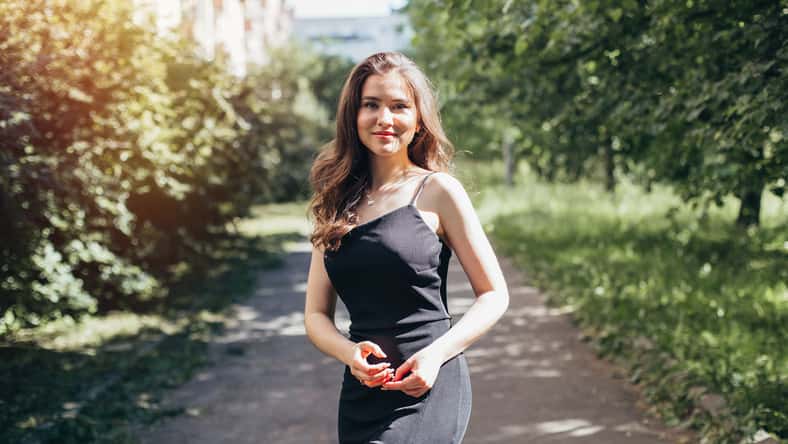 Young beautiful woman with long curly brunette hair in black dress, walking in summer park