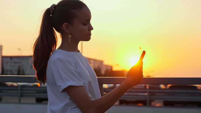 Schoolgirl looks into smartphone at sunset. Addicted young teenage girl with ponytail holds gadget in hand standing on bridge at back setting sun in summer